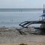 A traditional Filipino outrigger boat pulled up on a tidal flat at low tide, with people wading in the shallows beyond.