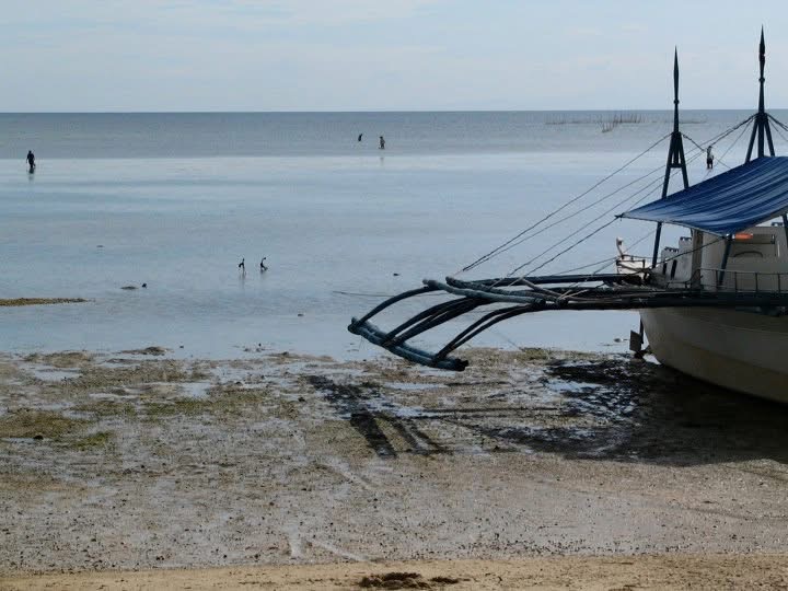 A traditional Filipino outrigger boat pulled up on a tidal flat at low tide, with people wading in the shallows beyond.